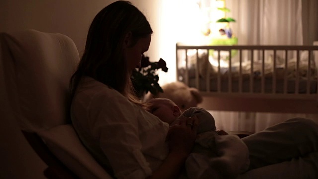 年轻美丽的母亲，在昏暗的灯光下给她的新生儿喂奶。妈妈母乳喂养婴儿。视频素材