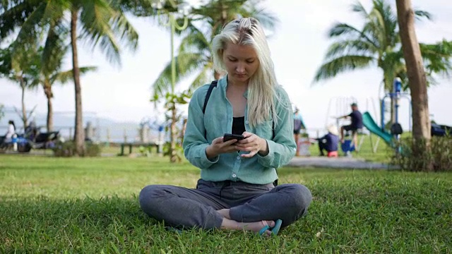 一位身穿绿色衬衫、一头金发的苗条美女坐在地上，用智能手机拍摄公园的背景。女孩在正方形触摸屏上微笑视频素材