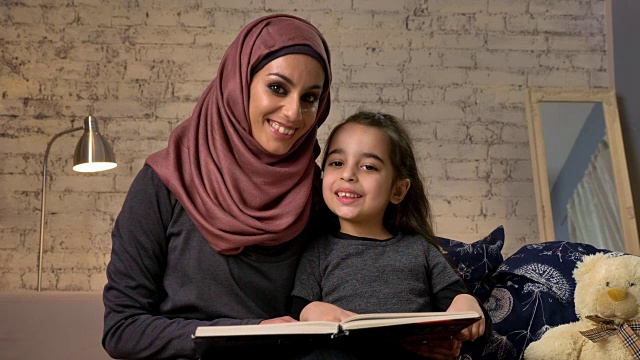 年轻女子戴着头巾微笑着和她的女儿坐在沙发上，教她如何阅读，书，幸福家庭的概念，近距离，家庭舒适的背景，50帧/秒视频下载