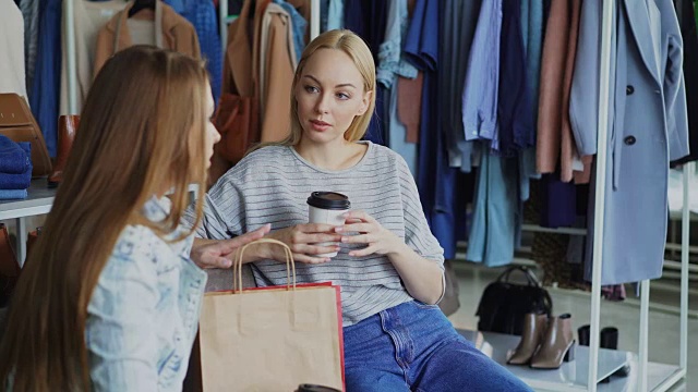 年轻女子坐在服装店，拿着咖啡和她的女性朋友聊天。女士们在购物后休息。视频素材