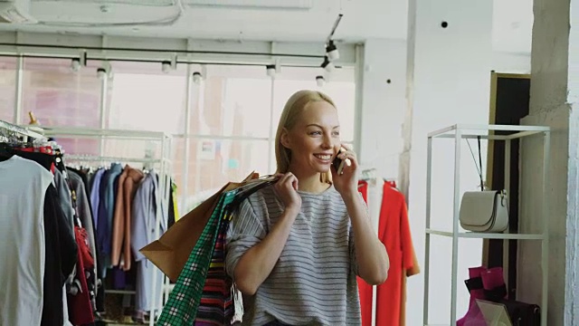 迷人的金发女人在大商店的货架和栏杆之间行走和讲手机。她提着包，微笑着看着周围时髦的衣服。视频素材