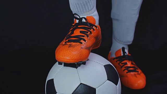 穿着靴子的足球运动员把他的腿放在一个球上，黑色的背景视频下载