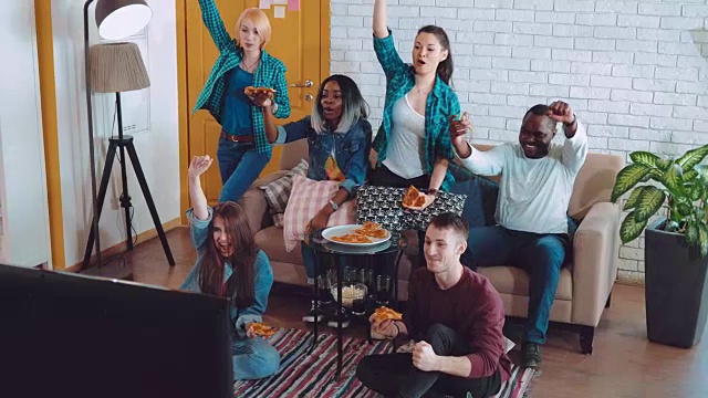 朋友们坐在一起看电视比赛，吃着披萨，讨论游戏，做手势。尖叫，站起来，举起他们的手，庆祝你最喜欢的球队的胜利视频素材