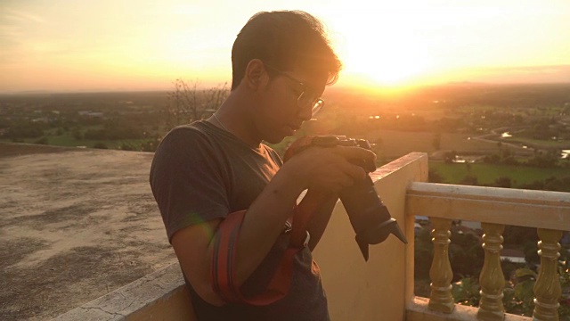日落时分，一个年轻的旅行者带着相机站在乡村小镇的山顶上视频下载