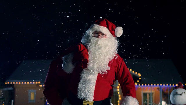 真正的圣诞老人肩扛红包，走出装饰着彩灯和花环的田园小屋。圣诞老人带来礼物和礼物。神奇的新年夜与飘落的雪。视频购买