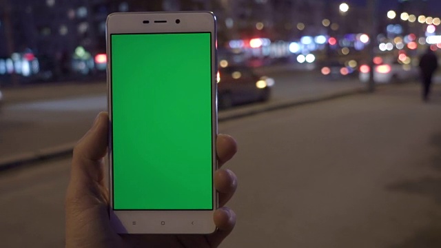 男子手握白色智能手机，在夜城灯光散景的背景下。绿色的屏幕。视频素材