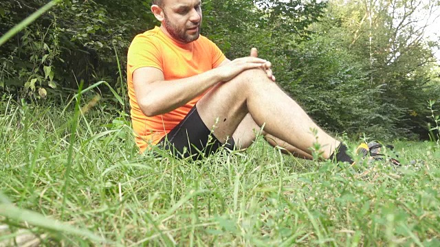 一名年轻人在慢跑时抽筋。停下来按摩膝盖。在夏日森林里，一个人跑过乡村。视频下载
