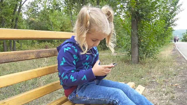 一个漂亮的小女孩坐在城市公园的长椅上使用智能手机上的应用程序。视频素材
