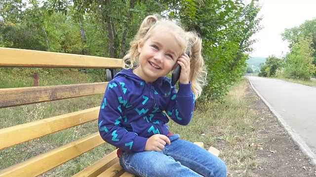 一个漂亮的小女孩坐在城市公园的长椅上打电话。视频素材