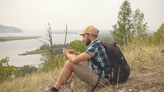 他坐在山里，正在使用智能手机。背着背包的人在树林里。徒步旅行。森林之旅露营。休闲。生态旅游。野外。冒险开始了。缓慢的运动。视频下载