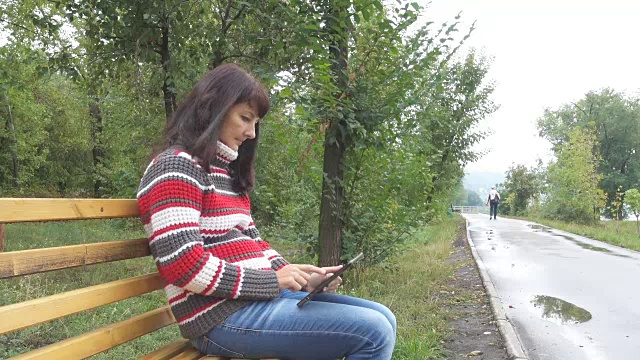 秋天的城市公园里，一名年轻女子拿着平板电脑坐在长椅上。女孩在他的平板电脑上玩游戏。视频素材