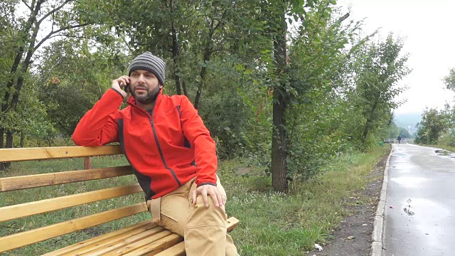 一个戴着帽子的男人坐在城市公园的长椅上打电话。视频素材