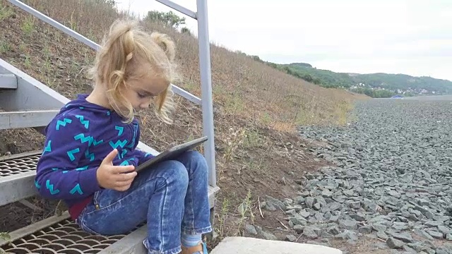一个漂亮的小女孩拿着平板电脑坐在河边的楼梯上。女孩在他的平板电脑上玩游戏。视频素材