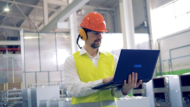 一个戴着安全帽的男人在一家工业工厂里用他的笔记本电脑工作。视频素材