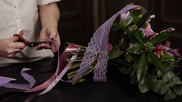 一个穿白衬衫的女孩剪下粉红色的长缎带和紫色的宽缎带来装饰桌上的一束花。近距离视频下载