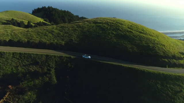 鸟瞰图的汽车驾驶下的乡村道路通过农村起伏的山与海洋的背景在日落视频素材