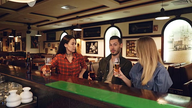 两位迷人的年轻女子和蓄着胡须的年轻男子坐在吧台边喝啤酒，相互碰杯聊天。朋友们聚在酒吧视频素材