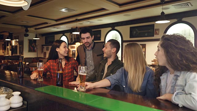 年轻人喝着啤酒在吧台边聊天，这时他们的几个朋友过来迎接他们。男人和女人互相问候，分享消息。视频素材
