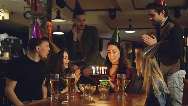 年轻漂亮的黑发女孩许愿，吹灭生日蛋糕上的蜡烛，与朋友在咖啡馆庆祝生日。快乐的人们戴着派对帽在鼓掌。视频下载