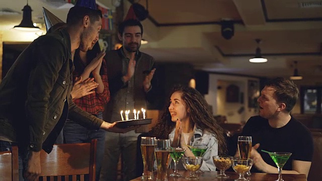 快乐的年轻人正把生日蛋糕带给坐在咖啡馆桌子旁闭着眼睛的年轻女士。她许愿，吹蜡烛，朋友们鼓掌。视频素材