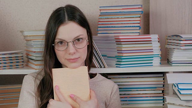 年轻的褐发女人戴眼镜，抱着一本书，看着相机，坐在书中。视频素材