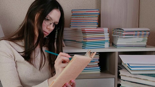 年轻的黑发女人戴着眼镜看书，用铅笔做笔记。视频素材