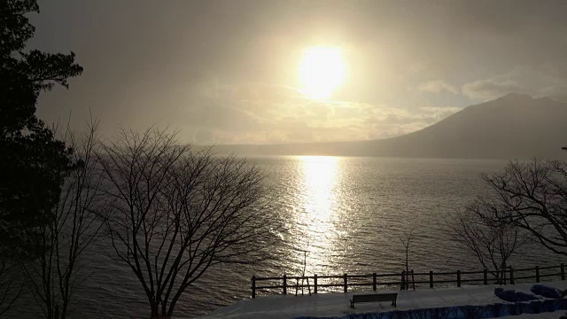 日出在波浪起伏的湖面上，冬天下雪，树木和长凳的剪影视频下载