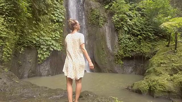 慢动作年轻女子享受新鲜瀑布在巴厘岛，印度尼西亚，亚洲。人们旅游乐趣自然的概念。女孩伸着胳膊视频下载