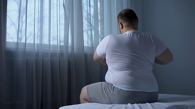 一个身形魁梧的胖子早上坐在床上伸展肌肉，感觉背部疼痛视频素材