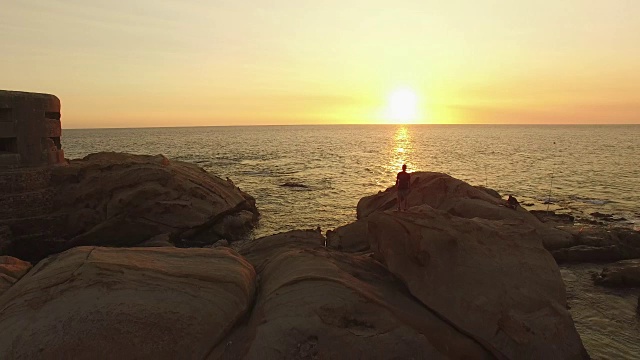 鸟瞰图，剪影男子在岩石顶部与手臂举起在日落的海滩视频素材