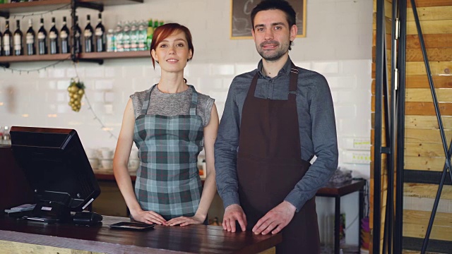 两个自信的服务员站在收银台的围裙在咖啡馆和微笑。成功的生意，快乐的人和餐饮的服务理念。视频素材