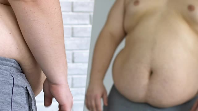 不开心的肥胖男性看着镜子里自己的大肚子，体重减轻，缺乏安全感视频素材