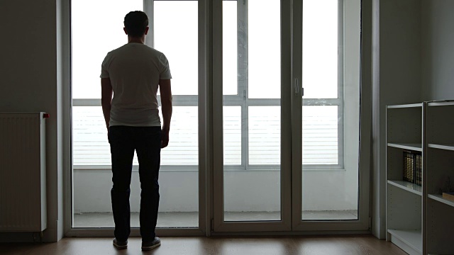 一个悲伤的年轻人的剪影站在家里的大窗户旁边视频素材