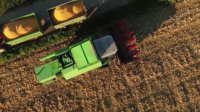 无人机视角联合收割机斜槽填充拖车收获的玉米在阳光明媚的农村田野，慢镜头视频素材