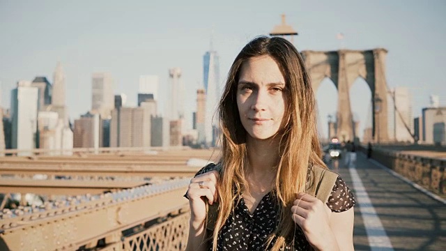 年轻积极的女性白人游客背包看相机，微笑在阳光灿烂的布鲁克林大桥，纽约4K视频素材