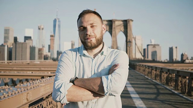 年轻的积极男性白人商人双臂交叉看着相机，微笑在布鲁克林大桥，纽约4K视频下载