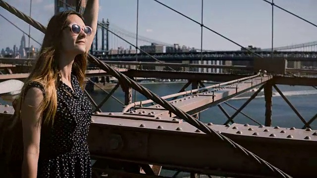 镜头跟随年轻快乐的女游客背包，戴着太阳镜沿着布鲁克林大桥走，欣赏4K风景视频素材