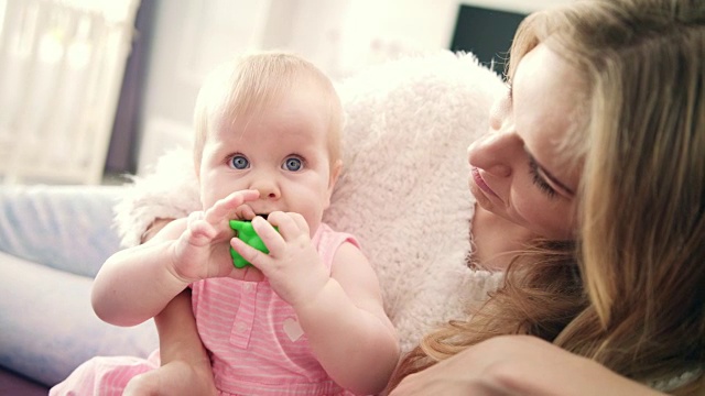 漂亮的女婴坐在妈妈的怀抱里。蹒跚学步的小女孩嘴里叼着玩具视频下载