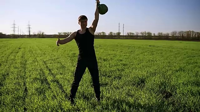 一个体格健壮的年轻人用一只手举起重物。美丽的大自然，站在绿色的草地上视频素材