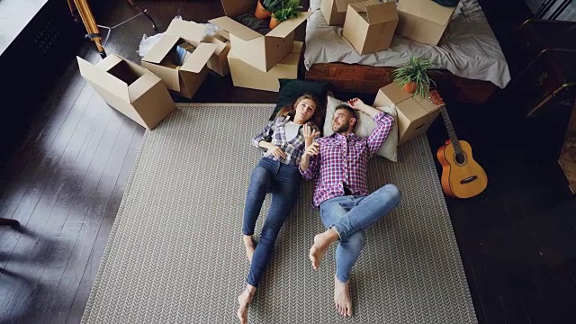 俯视图可爱的年轻夫妇躺在他们的新公寓卧室的地板上，交谈，亲吻和拥抱。纸箱、吉他、地毯和床都可以看到。视频素材