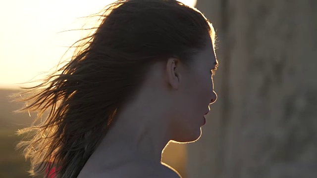 穿红衣服的年轻女子望着躲在悬崖后面的太阳，风吹散了她的头发视频素材