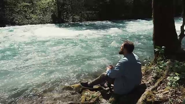 白天，一个留着胡子的男人独自坐在岸边，沉思地看着河流视频素材