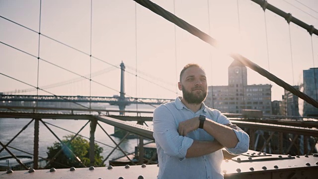 年轻快乐的白人男子站在布鲁克林大桥微笑，交叉双臂，看着周围享受4K的风景视频素材
