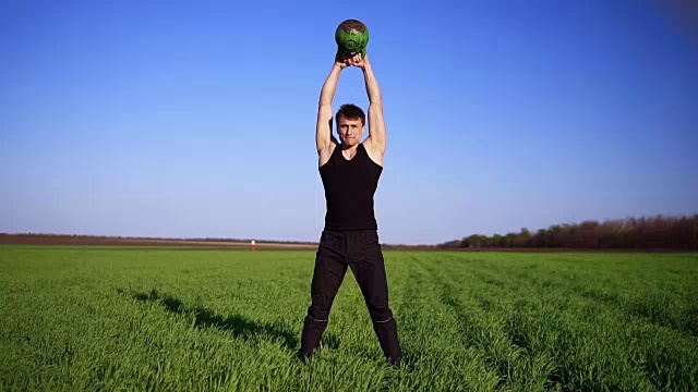 一个强壮的男人用重量演示一项运动。用两只手提起波波头。湛蓝的天空，绿色的田野。镜头前视频素材