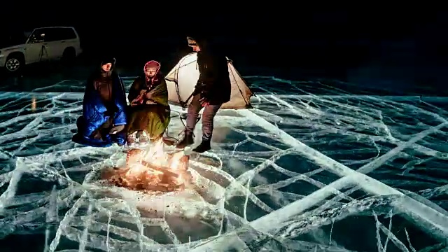 三个旅行者站在篝火旁，就在夜晚的冰面上。营地冰。帐篷紧挨着火。人们正在火炉旁取暖。延时与圆周运动。贝加尔湖。视频下载