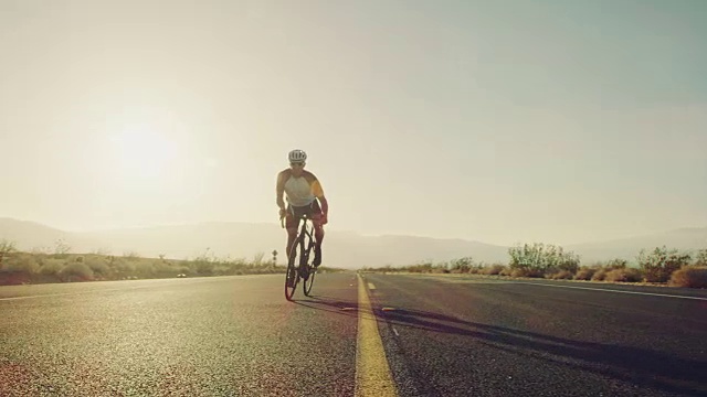 慢镜头近距离的男子骑在公路上的自行车在沙漠道路上在阳光明媚的一天与镜头光晕视频素材