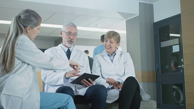 在医院大厅三位专业医生坐着讨论。他们使用平板电脑。繁忙的现代医院与最好的专家和医疗保健在国家。视频素材