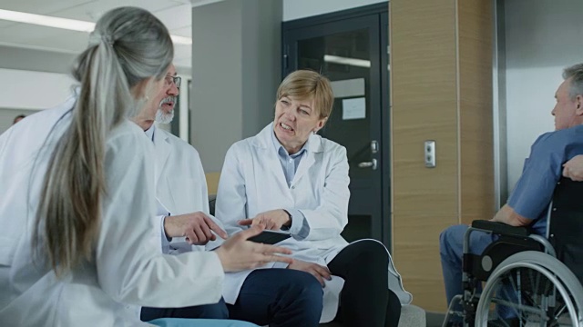 在医院大厅三位专业医生坐着讨论。他们使用平板电脑。繁忙的现代医院与最好的专家和医疗保健在国家。缓慢的运动。视频素材
