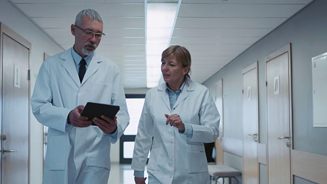 在医院里，两位医生走过医院的走廊，他们聊天并分享平板电脑。拥有病人和医务人员的现代化医院。视频素材