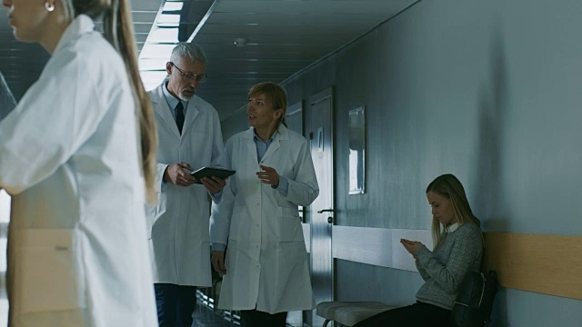 在医院里，两位医生走过医院的走廊，他们聊天并分享平板电脑。拥有病人和医务人员的现代化医院。缓慢的运动。视频素材